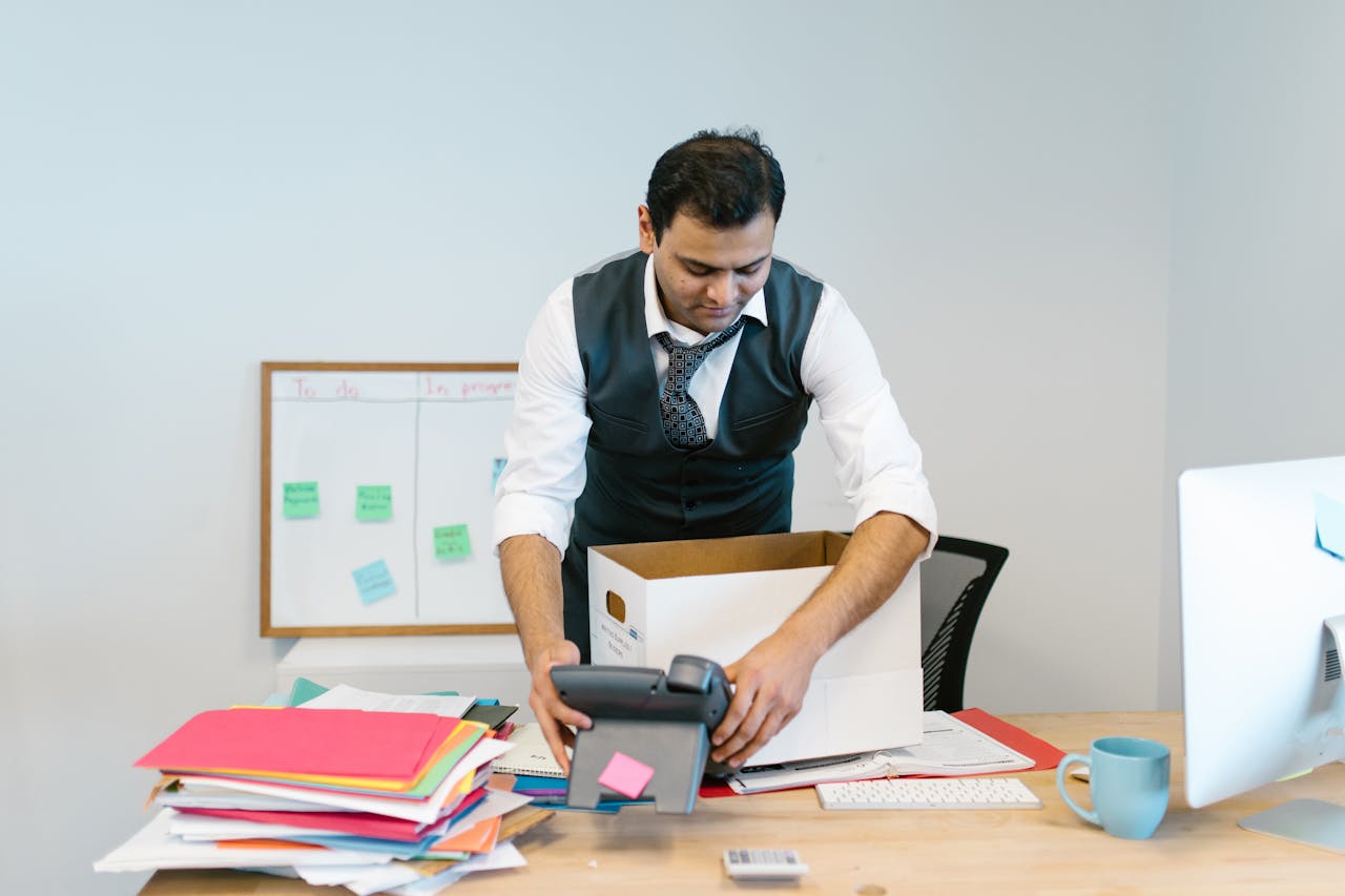 A man packing up his desk in an office setting with papers and a phone.