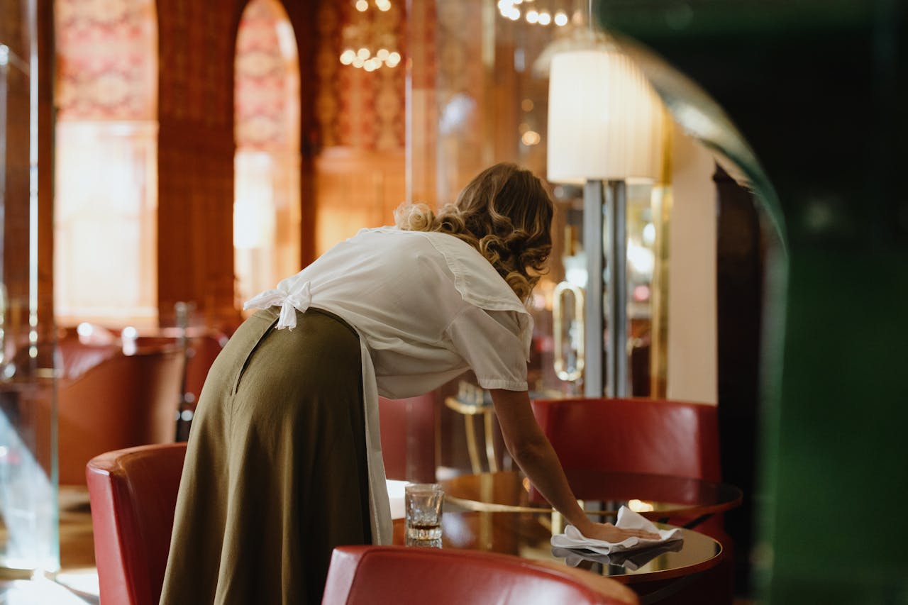 A woman is wiping a glass table in a cozy cafe with vintage decor and soft lighting.